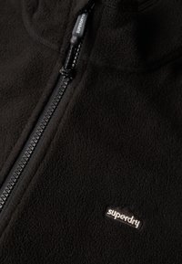Zwarte fleece jas met rits, met een opvallende zwarte rits en een klein wit "Superdry" logo aan de linkerkant. Zachte textuur.