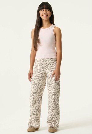 Fille aux longs cheveux noirs portant un haut sans manches rose clair, un pantalon beige à imprimé léopard et des chaussures beiges, debout et souriante.