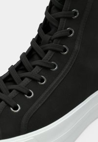 Μαύρο ψηλό αθλητικό παπούτσι με υφή υφάσματος, τυπικά κορδόνια, μεταλλικά ρουθούνια και λευκή λαστιχένια σόλα.