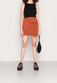 Mini-jupe orange avec détail de nœud, associée à un haut noir sans manches et des sandales noires épaisses, complétée par un sac noir texturé.