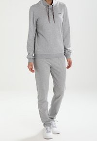 Ensemble de survêtement athlétique gris en tissu doux, avec capuche, cordons de serrage et coupe slim. Associé à des baskets blanches pour un look complet.