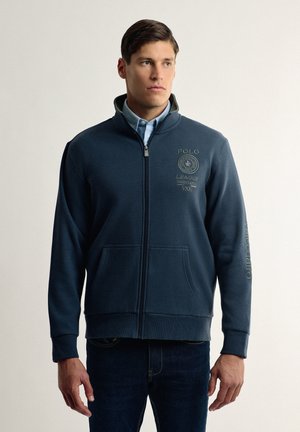 Sudadera de chándal azul marino con cremallera, hecha de material polar, que cuenta con un cuello alto, bolsillos laterales y logotipos bordados en dorado en el pecho y la manga.