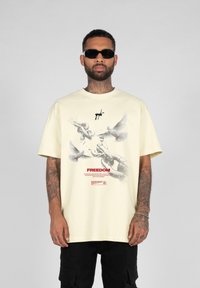 Cremefarvet bomulds T-shirt med et grafik af duer og kæder, med ordet "FREEDOM" i rød skrift. Løs pasform, korte ærmer.