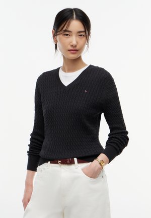 Kvinde iført en sort tekstureret V-hals sweater over en hvid skjorte med hvide bukser, et brunt bælte og et guld armbåndsur, hånd i lommen.
