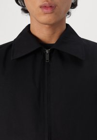 Črna jakna z zadrgo, ki ima koničast ovratnik, z diskretnim vzorcem in kovinsko zadrgo spredaj. Preprost in enobarven dizajn.