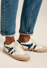 Bjeli niskotopni tenisice s naglascima u mornarsko plavoj boji i detaljima od sivoj antilopu, postavljene uz zarolane traperice. Jednostavni dizajn s vezanjem i gumeni potplat.