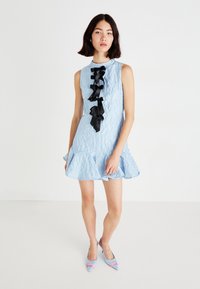 Abito azzurro chiaro, con tessuto texture, design senza maniche, orlo arricciato e dettagli in nastro nero lungo la parte frontale. Abbinato a tacchi azzurri chiari.