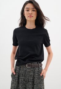 Maglione nero a maniche corte con colletto rotondo, abbinato a una gonna a pieghe in pattern houndstooth. Cintura marrone con fibbia rettangolare in argento.