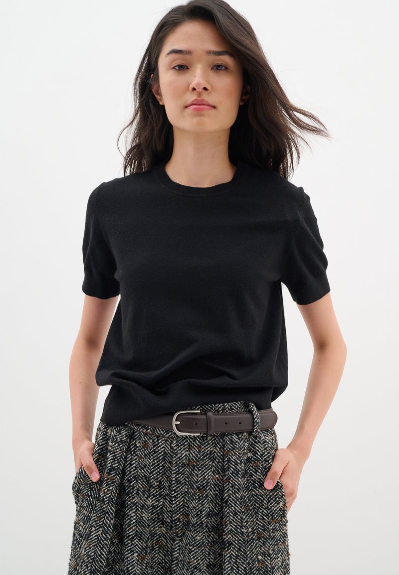 Maglione nero a maniche corte con colletto rotondo, abbinato a una gonna a pieghe in pattern houndstooth. Cintura marrone con fibbia rettangolare in argento.