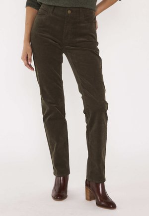 Persoon die donkergroene corduroy broek en bruine enkellaarsjes met hakken draagt, staand tegen een effen achtergrond.
