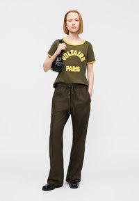 Ελιά πράσινο t-shirt με κίτρινη διακόσμηση και εκτύπωση "VOLTAIRE PARIS", συνδυασμένο με φαρδιά, σκουρόχρωμα καφέ παντελόνια και μαύρα παπούτσια, κρατώντας μια μικρή τσάντα.