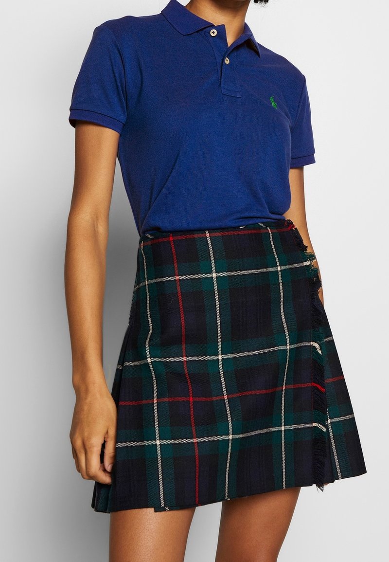 Personne portant un polo bleu à manches courtes et une mini-jupe à carreaux vert foncé, bleu marine, rouge et blanc avec des bords effilochés.