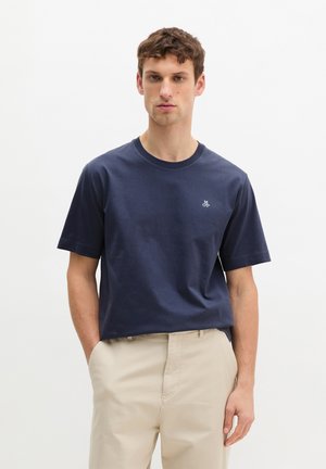 Jonge man staat met zijn handen in de zakken van een beige broek, draagt een navy blauw T-shirt met korte mouwen en een klein wit logo op de borst.
