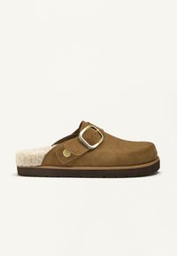 Clog-uri din suede maro cu o căptușeală pufoasă din blană albă, cataramă metalică ajustabilă și o talpă groasă din cauciuc cu un model de profil.