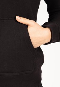 Zwarte sweatshirt met een voorzak, met een close-up van een hand die in de zak rust. Getextureerde stof met een gladde afwerking.