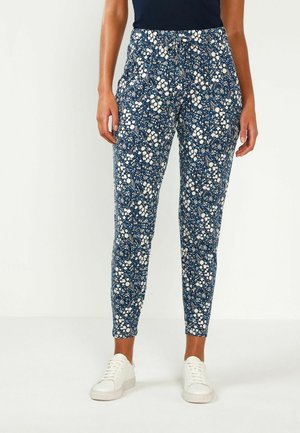 Femme portant un pantalon slim bleu marine à motif floral avec des baskets blanches, debout devant un fond clair uni.