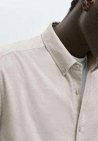 Camicia button-up beige con una texture morbida, caratterizzata da un colletto e cuciture visibili. Uno strato interno bianco è parzialmente esposto all'area del collo.