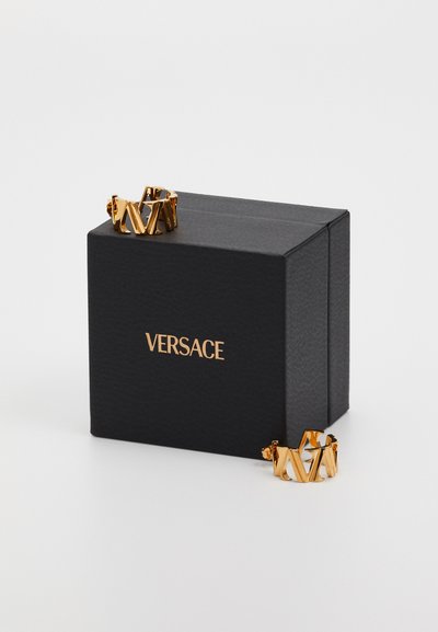 Bague en ton or avec un design géométrique, présentant des formes entrelacées. Présentée dans une boîte texturée noire de Versace avec un logo doré.