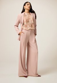Blush roze kostuum met een getailleerde blazer en wijde broekspijpen. Doorzichtige bloes met bloemen eronder. Geaccentueerd met gouden oorbellen en puntige hakken.