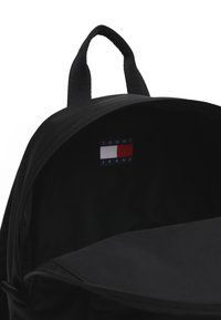 Mochila negra con una textura suave, cierre de cremallera y asa en la parte superior. El interior presenta un parche con el logo en rojo, blanco y azul.