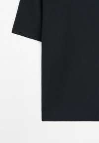 Tissu tricoté texturé noir à manches courtes avec un motif alvéolé serré, montrant un ourlet de chemise ou de pull et le bord de la manche.