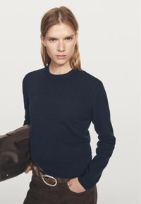 Granatowy sweter dziany o okrągłym dekolcie i długich rękawach, zestawiony z brązowymi spodniami i brązową torebką. Gładka faktura, minimalistyczny design.