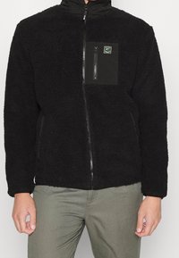 Chaqueta de forro polar negra con exterior texturizado, cremallera de largo completo y un bolsillo en el pecho con cremallera negra que presenta un parche con logo verde.
