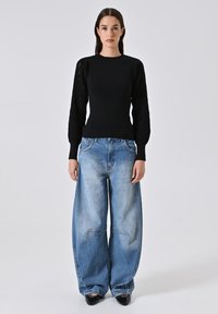 Schwarzer Strickpullover mit strukturierten Puffärmeln, kombiniert mit übergroßen hellblauen Jeans, die gerade Beine und leichte Ausbleichungen aufweisen.
