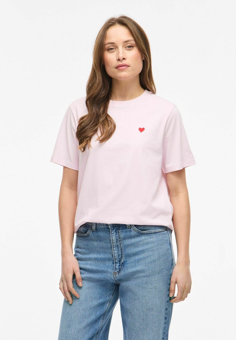 Femme aux longs cheveux bruns portant un t-shirt rose clair avec un petit cœur rouge sur la poitrine et un jean bleu, debout devant un fond blanc.