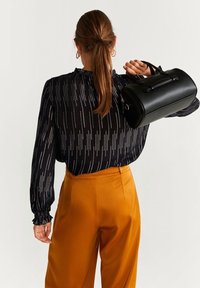 Mujer con coleta castaña, vistiendo blusa negra con estampado y pantalones mostaza, sosteniendo un bolso negro en el hombro, de pie contra un fondo blanco.