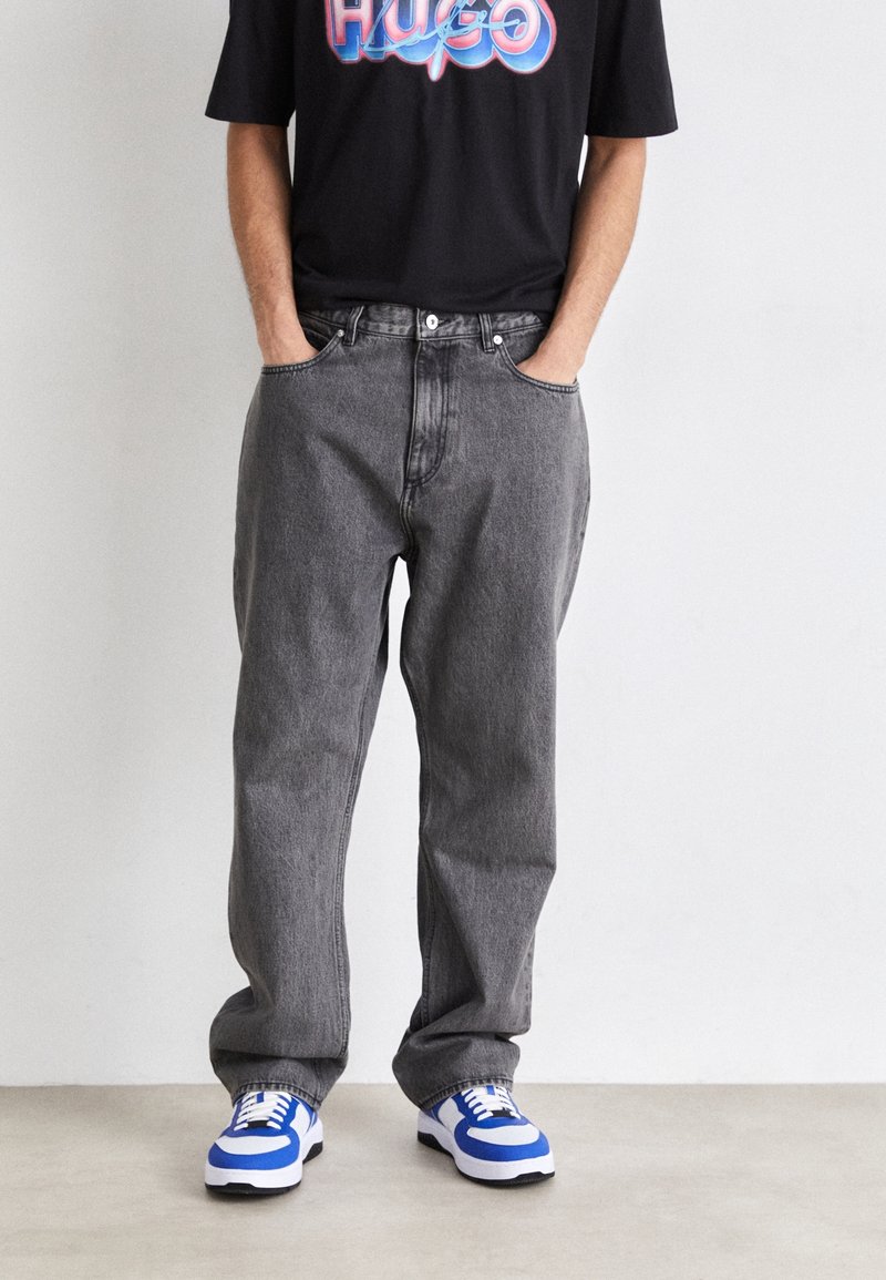 Grå denimjeans med avslappnad passform, med fickor framtill och knappstängning. Matchas med blå och vita sneakers och en svart t-shirt.