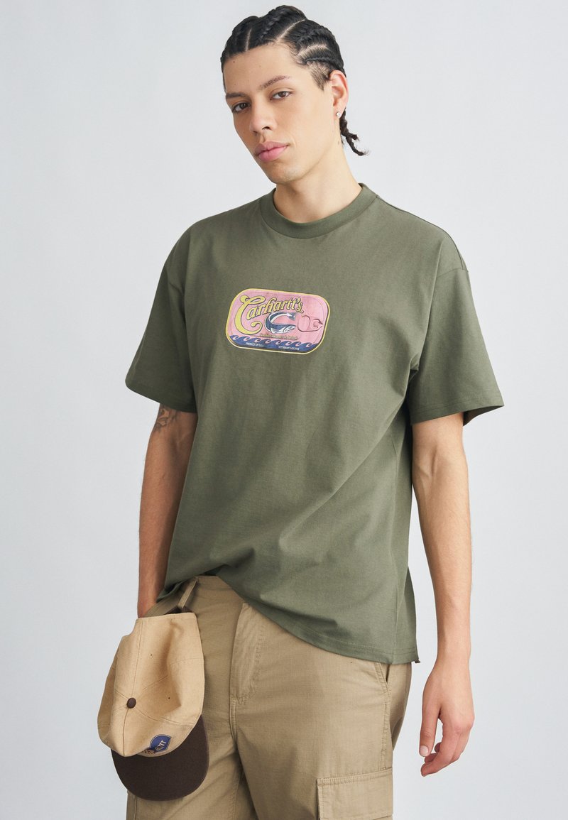 Olívazöld rövid ujjú pamut póló színes, grafikával díszített mintával a mellrészen. Bézs cargo rövidnadrággal és bézs sapkával viselve.