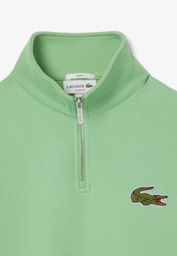 Suéter de color verde claro con un alto cuello y cierre de media cremallera. Presenta un logo de cocodrilo bordado en el pecho. Textura suave y ajuste holgado.