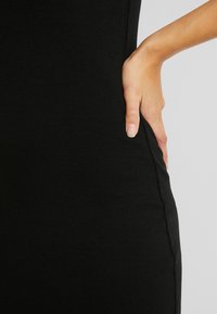 Handen vilar på höften, iförd ett åtsittande svart ribbat plagg som visar sid sömnens detalj mot en enkel ljus bakgrund.