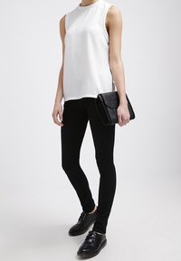 Top blanco sin mangas con tejido texturizado, combinado con jeans ajustados negros y zapatos de cordones negros. Cartera negra sostenida en la mano derecha.