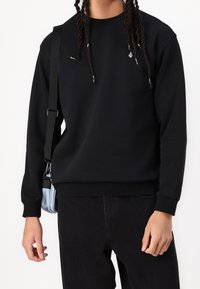 Czarny sweatshirt z okrągłym dekoltem wykonany z bawełny, z prążkowanymi mankietami i wykończeniem, z małym detalem logotypu. Zestawiony z czarnymi spodniami i torbą na ramię.