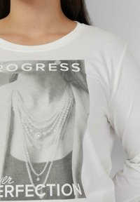 Witte lange mouwen shirt met een zwart-witte afbeelding met tekst, versierd met strass-steentjes die laagjes parelkettingen nabootsen.