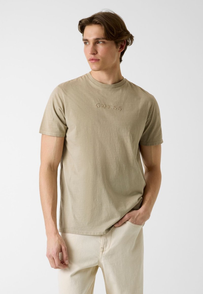 Jeune homme portant un t-shirt beige de la marque Guess et un pantalon clair, debout avec une main dans la poche, regardant vers sa gauche sur un fond clair uni.