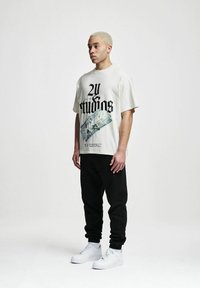 Kreminė grafika t-shirt su juodu tekstu ir pinigų spauda, derinama su juodomis kelnėmis ir baltais sportbačiais.