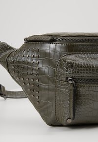 Banane en cuir synthétique effet crocodile vert olive avec poche zippée sur le devant, détails métalliques cloutés et bandoulière réglable.