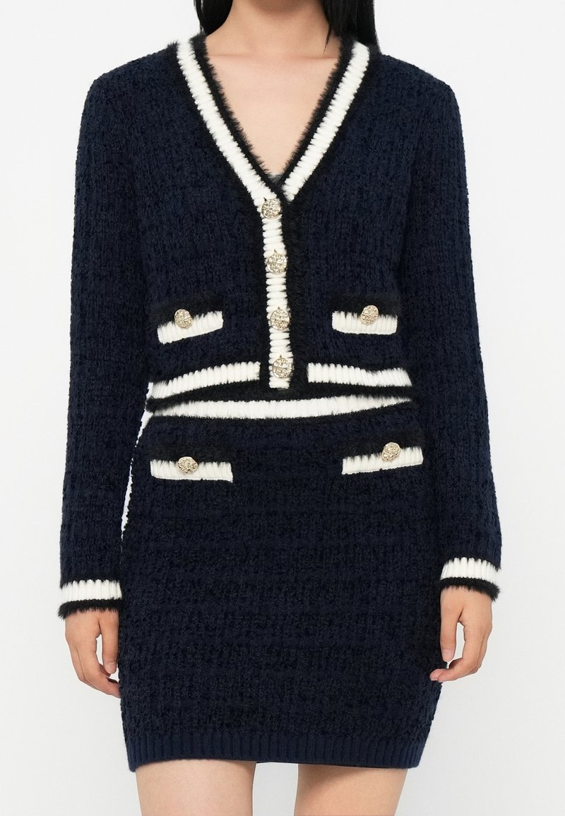 Femme portant un ensemble cardigan et jupe en maille texturée bleu marine avec des bordures blanches et des boutons dorés aux poignets, poches et devant.
