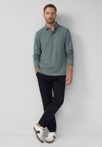Homme portant un polo vert à manches longues, un pantalon noir et des baskets blanches, se tenant les jambes croisées contre un mur gris uni.