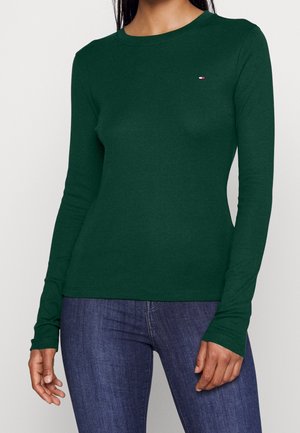 Nainen pukeutuneena istuvaan tummanvihreään pitkähihainen yläosaan, jossa on pieni punainen, valkoinen ja sininen logo vasemmassa rinnassa, ja sinisiin farkkuihin.