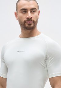T-shirt de sport blanc à manches courtes, de coupe ajustée, en tissu lisse et extensible, avec le logo "Champion" gris sur la poitrine.