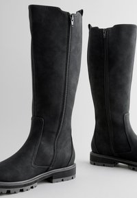 Paar hohe schwarze Stiefel mit seitlichen Reißverschlüssen und groben Sohlen, aufrecht auf hellem Hintergrund dargestellt.