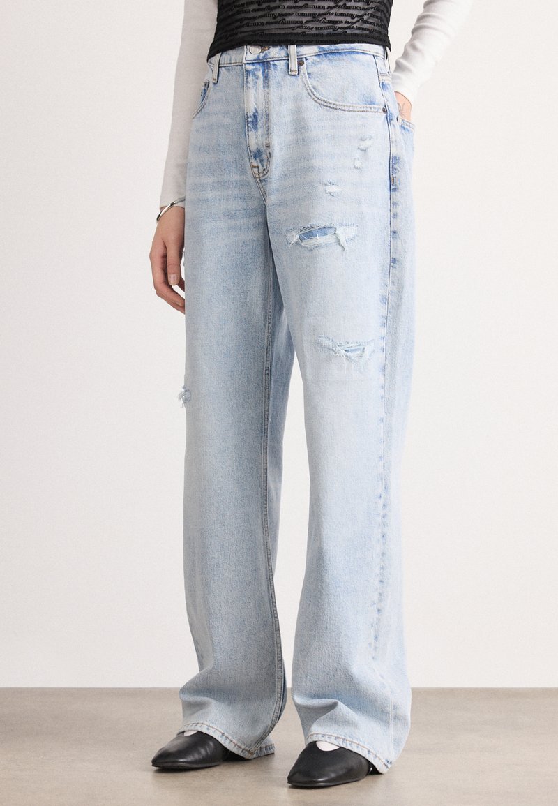 Los jeans de mezclilla anchos de color azul claro presentan detalles desgastados, una cintura alta y bajos deshilachados. La modelo los combina con zapatos negros.