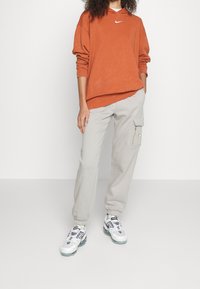 Felpa arancione Nike con logo bianco, abbinata a pantaloni cargo grigi chiari con una tasca laterale e sneakers bianche con dettagli grigi.