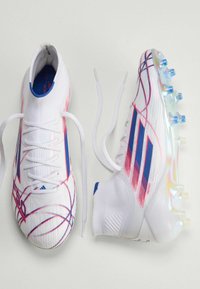 Ζευγάρι λευκών ποδοσφαιρικών παπουτσιών με μπλε και κόκκινες λεπτομέρειες, κορδόνια ξεχειλωμένα, σε θέα από πάνω και από το πλάι σε απλό φόντο.