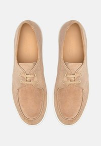 Bež nubuk tenisice s okruglim vrhom, minimalnim vezanjem i bijelim gumeni potplat. Glatka tekstura s blagim sjajem i čistim detaljima šivanja.