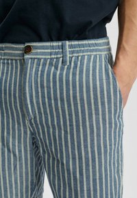 Gestreepte blauw en crème broek met een visgraatpatroon, voorzien van een knoopsluiting, riemlussen en zijzakken. De stof is geweven.
